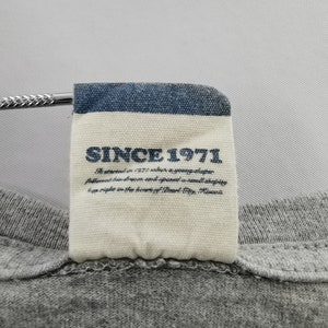 May include: Close-up of a gray t-shirt with a tag. The tag has a blue border and the text "SINCE 1971" in navy blue. Below the text is a description of the brand's history.