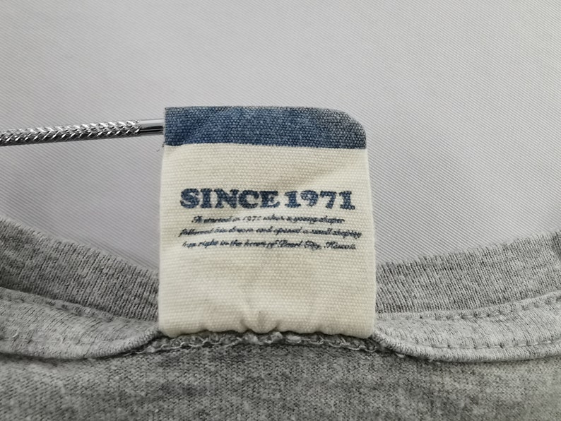 May include: Close-up of a gray t-shirt with a tag. The tag has a blue border and the text "SINCE 1971" in navy blue. Below the text is a description of the brand's history.
