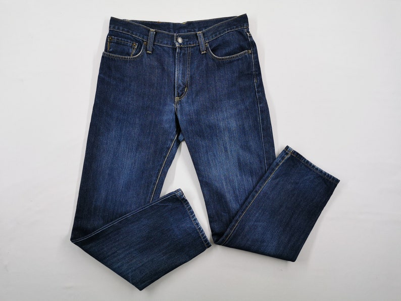 May include: Dark blue denim jeans with a classic five-pocket design. The jeans are laid flat, showcasing the front and back pockets, belt loops, and button closure. The fabric has a slightly faded appearance.