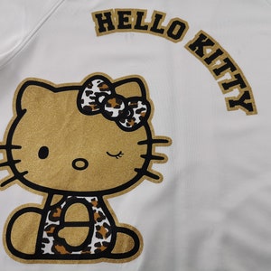 May include: White sweatshirt featuring a gold glitter Hello Kitty design. The design includes a winking Hello Kitty with a leopard print bow and accents. The words "HELLO KITTY" are arched above the character in gold.