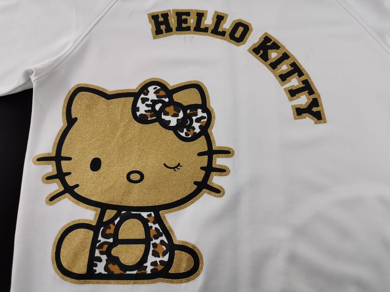 May include: White sweatshirt featuring a gold glitter Hello Kitty design. The design includes a winking Hello Kitty with a leopard print bow and accents. The words "HELLO KITTY" are arched above the character in gold.