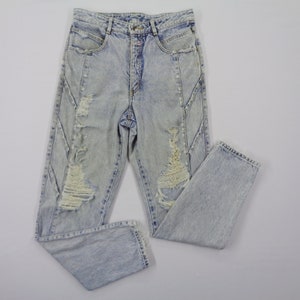 May include: Light blue acid wash jeans with distressed details. The jeans feature a high waist, a button and zipper closure, and ripped sections on the front of the legs. The jeans are folded on a white surface.