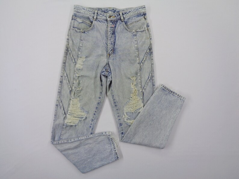 May include: Light blue acid wash jeans with distressed details. The jeans feature a high waist, a button and zipper closure, and ripped sections on the front of the legs. The jeans are folded on a white surface.