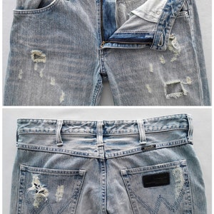 Pu&ograve; includere: Jeans in denim blu chiaro con dettagli strappati, tra cui aree strappate. I jeans hanno una chiusura con bottone e cerniera, tasche anteriori e posteriori, e l'etichetta del marchio "Wrangler" &egrave; visibile. Stile classico.