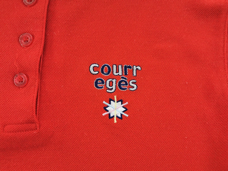 K&ouml;nnte beinhalten: Ein rotes Poloshirt mit drei transparenten Kn&ouml;pfen und einem Kragen. Auf dem Shirt ist der Text "COURR eges" in Wei&szlig; und Marineblau sowie ein kleines, farbenfrohes Emblem gestickt.