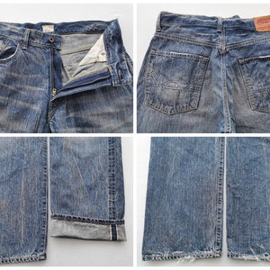 May include: Blue denim jeans with a faded wash. The jeans have a button and zipper closure, front and back pockets, and a leather patch on the back. The cuffs are rolled up, revealing a red selvedge detail. The jeans are a classic, casual style.
