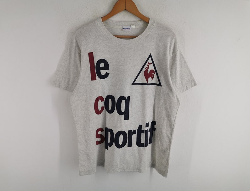 May include: A light grey t-shirt featuring the text "le coq sportif" in a bold font. The word "le" is in a dark red colour, "coq" is in a burgundy colour, and "sportif" is in a dark blue colour. A red rooster logo is inside a black triangle.