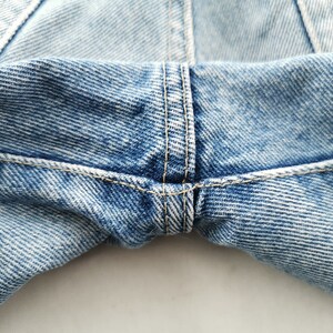 May include: Close-up of light blue denim jeans. The image highlights the stitching and construction details, including the seams and the fabric's texture. The jeans appear to be well-worn, with a faded appearance.