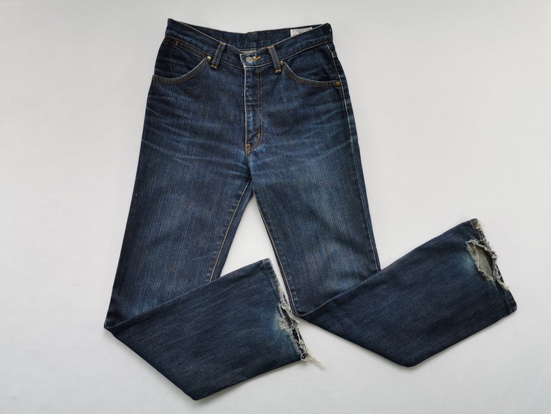 May include: Dark wash denim jeans with a classic five-pocket design. The jeans have a button and zip closure, and a slight flare at the hem. The denim has a worn look with some distressing at the bottom of the legs.