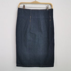Puede incluir: Una falda lápiz de mezclilla azul oscuro con diseño de cintura alta, detalles de costuras verticales y pespuntes en contraste. La falda cuelga de una percha de madera con clips metálicos, mostrando una silueta recta y un lavado índigo.