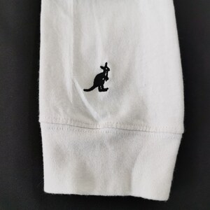 Op de afbeelding: Witte lange mouwen shirtmouw met een zwart kangoeroe logo geborduurd op de linkerbovenkant. De manchet is geribbeld en de stof lijkt een zachte katoenmix te zijn. Het kangoeroe logo is een eenvoudig, gestileerd ontwerp.
