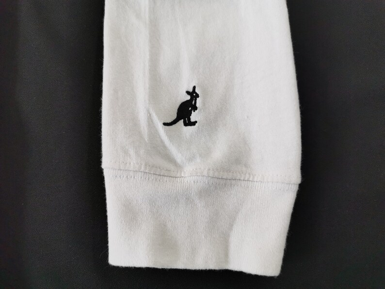 Op de afbeelding: Witte lange mouwen shirtmouw met een zwart kangoeroe logo geborduurd op de linkerbovenkant. De manchet is geribbeld en de stof lijkt een zachte katoenmix te zijn. Het kangoeroe logo is een eenvoudig, gestileerd ontwerp.
