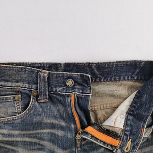 Op de afbeelding: Close-up van een blauwe spijkerbroek met een open gulp en een oranje rits. De jeans heeft een knoopsluiting en een klein voorzakje. De denim heeft een verweerde uitstraling.