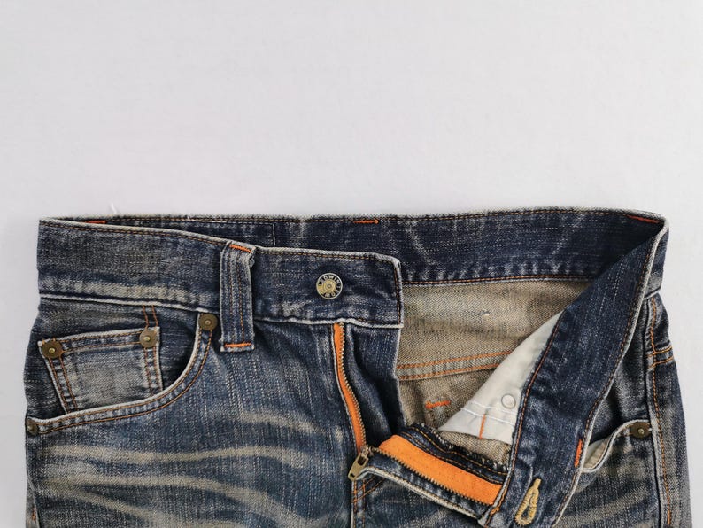Op de afbeelding: Close-up van een blauwe spijkerbroek met een open gulp en een oranje rits. De jeans heeft een knoopsluiting en een klein voorzakje. De denim heeft een verweerde uitstraling.