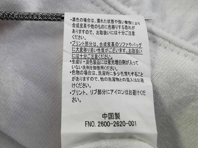 Pu&ograve; includere: Un'etichetta di tessuto bianco con testo giapponese e le parole "中国製" (Prodotto in Cina). L'etichetta include anche il numero "FNO. 2600-2620-001". &Egrave; visibile un ago d'argento.