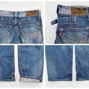 May include: Blue denim jeans with a faded wash, featuring a unique back design with a belt loop detail. The jeans have decorative stitching on the back pockets and a red tag. The label reads "Blue Trip".