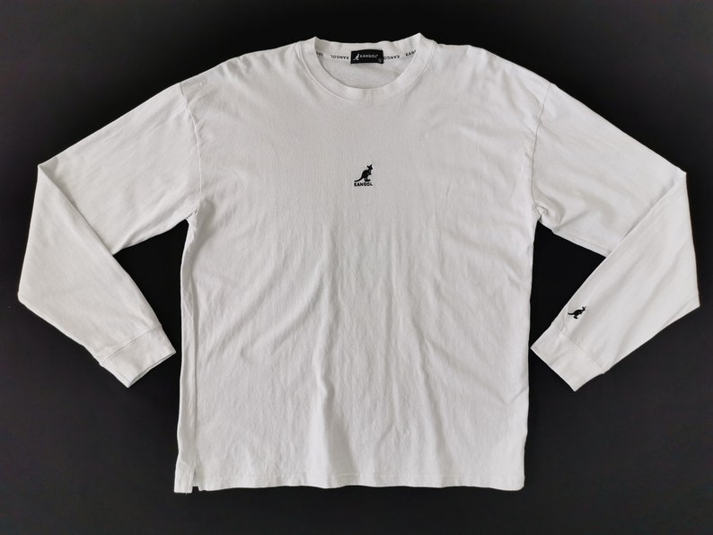 Op de afbeelding: Wit shirt met lange mouwen en een ronde hals. Het shirt heeft een klein zwart kangoeroe-logo op de borst en een bijpassend logo op de linker manchet. De merknaam is in het zwart rond de kraag gedrukt.