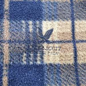 Puede incluir: Primer plano de una tela de forro polar a cuadros azul, beige y blanco. La tela tiene un aspecto suave y texturizado. Se ve un logotipo bordado de un p&aacute;jaro y las palabras "SCOTT COLLECTION".