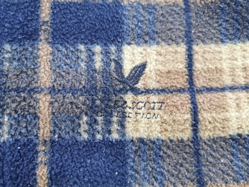 Puede incluir: Primer plano de una tela de forro polar a cuadros azul, beige y blanco. La tela tiene un aspecto suave y texturizado. Se ve un logotipo bordado de un p&aacute;jaro y las palabras "SCOTT COLLECTION".