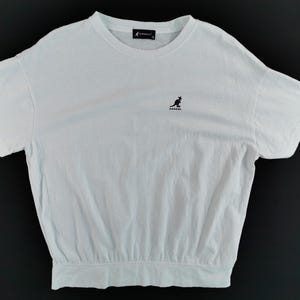 Pu&ograve; includere: T-shirt bianco con girocollo e maniche corte. Presenta un piccolo logo di canguro nero sul petto e un'etichetta nera sul retro del collo. La maglietta ha un orlo arricciato.