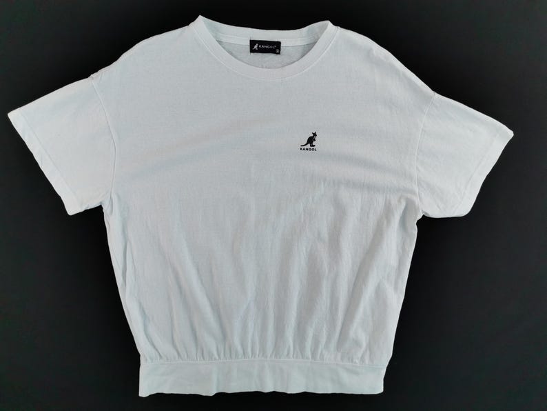 Pu&ograve; includere: T-shirt bianco con girocollo e maniche corte. Presenta un piccolo logo di canguro nero sul petto e un'etichetta nera sul retro del collo. La maglietta ha un orlo arricciato.