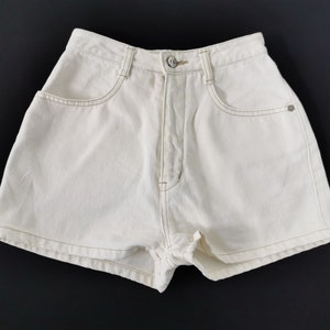 Puede incluir: Pantalones cortos vaqueros color crema con cintura alta y diseño clásico de cinco bolsillos. Los pantalones cortos tienen cierre de botón y costuras visibles en un color marrón claro contrastante. La prenda está hecha de algodón resistente.