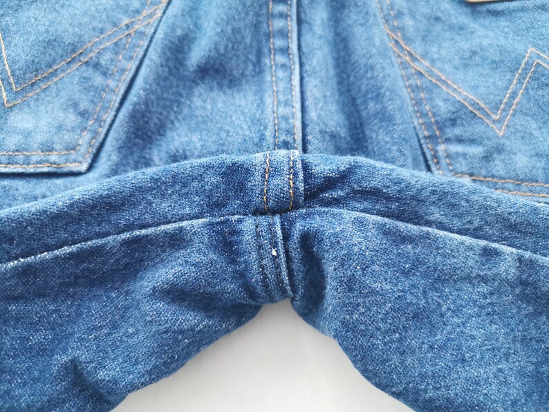 K&ouml;nnte beinhalten: Nahaufnahme einer blauen Jeans. Das Bild zeigt die Ges&auml;&szlig;taschen, N&auml;hte und den Taillenbereich. Die N&auml;hte sind in einer kontrastierenden beigen Farbe gehalten. Der Denim hat einen klassischen, abgenutzten Look.