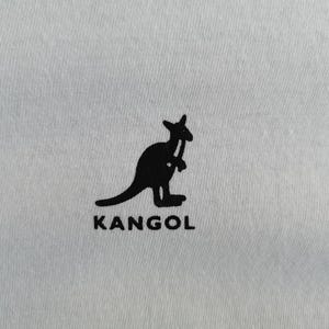 Pu&ograve; includere: Primo piano di un tessuto grigio chiaro con una silhouette nera di un canguro che tiene un bastone da passeggio. La parola "KANGOL" &egrave; stampata in nero sotto.