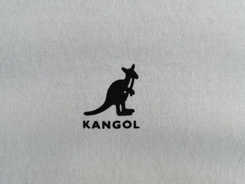 Pu&ograve; includere: Primo piano di un tessuto grigio chiaro con una silhouette nera di un canguro che tiene un bastone da passeggio. La parola "KANGOL" &egrave; stampata in nero sotto.