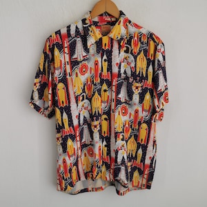 May include: Short-sleeved shirt with a space-themed print. The design includes rockets, astronauts, and planets in red, yellow, and white on a dark blue background. The shirt is displayed on a wooden hanger.