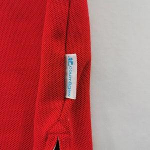 K&ouml;nnte beinhalten: Nahaufnahme eines roten Poloshirts mit strukturiertem Strickstoff. Ein wei&szlig;es Etikett mit dem blauen Text "courr&egrave;ges" ist an der Seite des Shirts angen&auml;ht. Das Shirt hat einen klassischen Kragen und eine Knopfleiste.