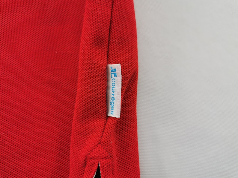 K&ouml;nnte beinhalten: Nahaufnahme eines roten Poloshirts mit strukturiertem Strickstoff. Ein wei&szlig;es Etikett mit dem blauen Text "courr&egrave;ges" ist an der Seite des Shirts angen&auml;ht. Das Shirt hat einen klassischen Kragen und eine Knopfleiste.