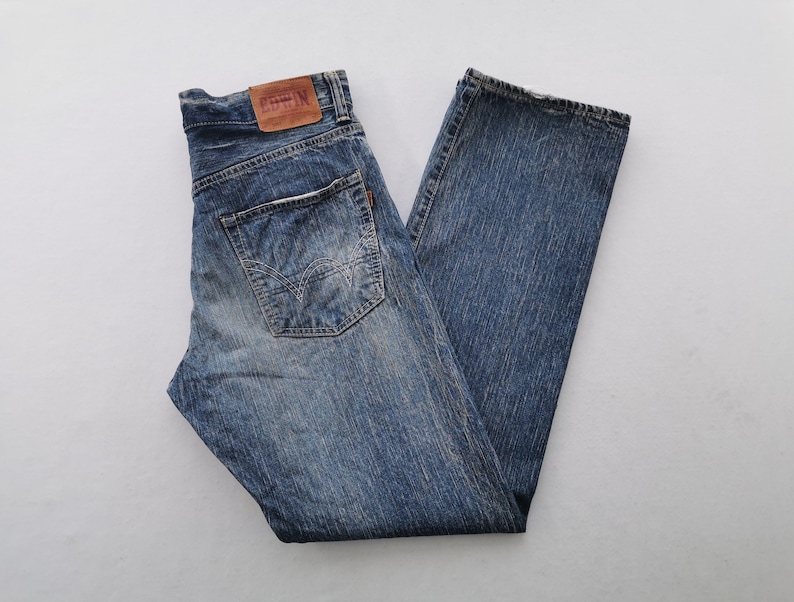 May include: Blue denim jeans with a faded wash. The jeans have a classic five-pocket design, a brown leather patch, and a straight leg. The jeans are folded neatly on a white surface.