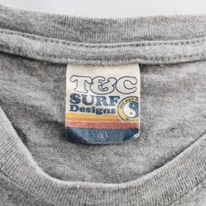 May include: Close-up of a gray t-shirt with a tag that reads "T&C SURF Designs." The tag features a retro design with a sunset-colored stripe and a yin-yang symbol. The shirt is a size large.