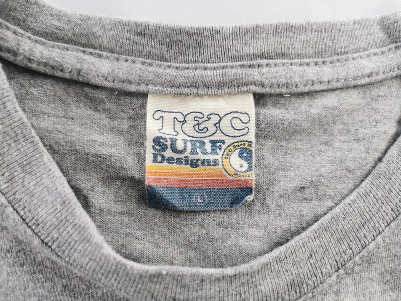 May include: Close-up of a gray t-shirt with a tag that reads "T&C SURF Designs." The tag features a retro design with a sunset-colored stripe and a yin-yang symbol. The shirt is a size large.