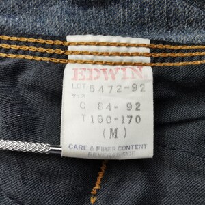 May include: Close-up of a denim garment with a white tag. The tag features the brand name "EDWIN" in red, along with product details and size information. The denim has visible stitching and a textured appearance.