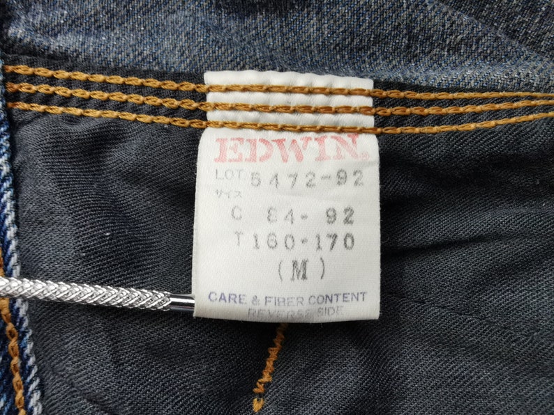 May include: Close-up of a denim garment with a white tag. The tag features the brand name "EDWIN" in red, along with product details and size information. The denim has visible stitching and a textured appearance.