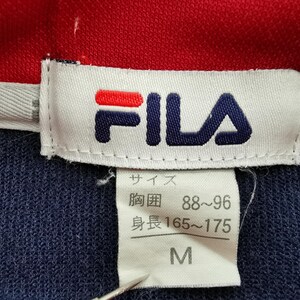 Pu&ograve; includere: Primo piano di un'etichetta di camicia Fila blu navy e rossa. L'etichetta mostra il logo Fila in rosso e blu navy. L'etichetta include anche informazioni sulla taglia in giapponese, con la taglia indicata come M.