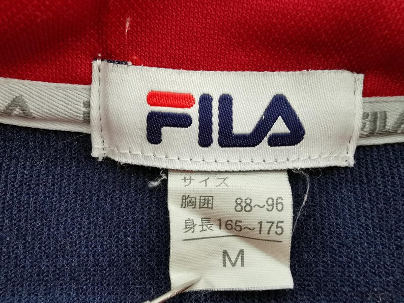 Pu&ograve; includere: Primo piano di un'etichetta di camicia Fila blu navy e rossa. L'etichetta mostra il logo Fila in rosso e blu navy. L'etichetta include anche informazioni sulla taglia in giapponese, con la taglia indicata come M.