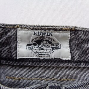 Puede incluir: Primer plano de unos vaqueros grises. La etiqueta blanca dice "EDWIN INTERNATIONAL BASIC" con un gr&aacute;fico de globo. Los vaqueros tienen un aspecto texturizado y costuras visibles.