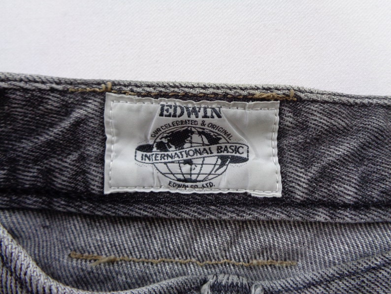 Puede incluir: Primer plano de unos vaqueros grises. La etiqueta blanca dice "EDWIN INTERNATIONAL BASIC" con un gr&aacute;fico de globo. Los vaqueros tienen un aspecto texturizado y costuras visibles.