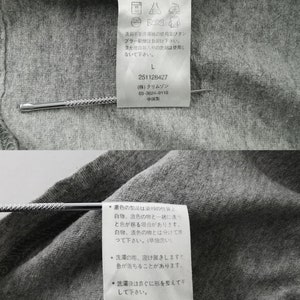 May include: Close-up of a gray cotton garment tag with Japanese text and care symbols. The tag includes washing instructions, size "L", and the number "251128427". A silver pin is inserted through the tag.