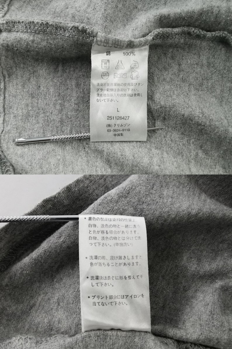 May include: Close-up of a gray cotton garment tag with Japanese text and care symbols. The tag includes washing instructions, size "L", and the number "251128427". A silver pin is inserted through the tag.