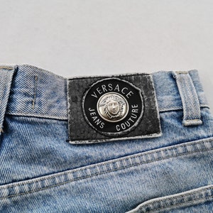 May include: Close-up of light blue denim jeans. The back of the jeans features a black leather patch with the words "VERSACE JEANS COUTURE" around a silver Medusa head emblem. The jeans have visible stitching.
