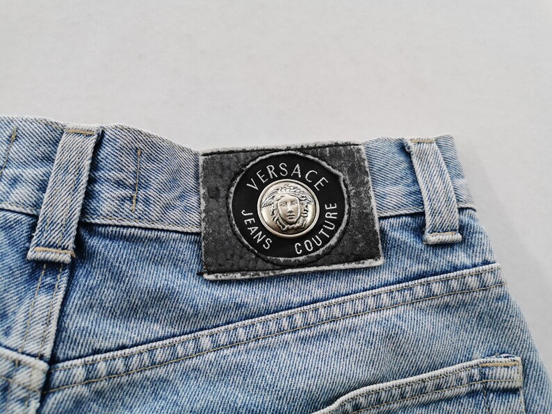 May include: Close-up of light blue denim jeans. The back of the jeans features a black leather patch with the words "VERSACE JEANS COUTURE" around a silver Medusa head emblem. The jeans have visible stitching.
