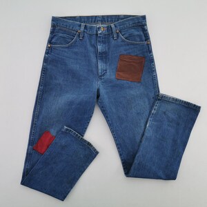May include: Blue denim jeans featuring a high-waisted cut with a button fly closure. The front of the jeans displays a large rectangular brown patch on the right thigh and a smaller rectangular red patch on the lower left leg near the hem.