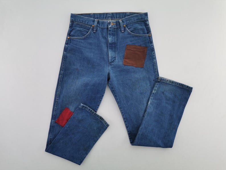 May include: Blue denim jeans featuring a high-waisted cut with a button fly closure. The front of the jeans displays a large rectangular brown patch on the right thigh and a smaller rectangular red patch on the lower left leg near the hem.