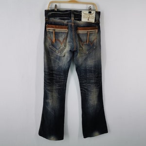 Pu&ograve; includere: Jeans blu in denim con lavaggio effetto used, con dettagli in pelle marrone sulle tasche posteriori e una cerniera. I jeans hanno un'apertura della gamba svasata e sono appesi a una gruccia di legno.