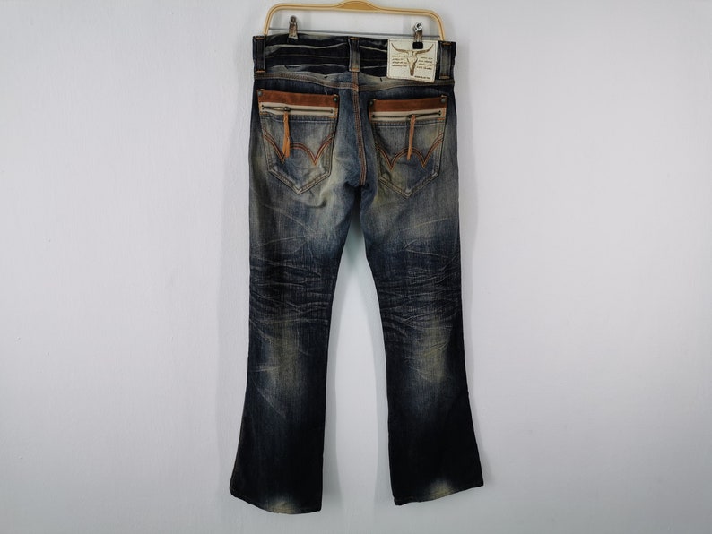 Pu&ograve; includere: Jeans blu in denim con lavaggio effetto used, con dettagli in pelle marrone sulle tasche posteriori e una cerniera. I jeans hanno un'apertura della gamba svasata e sono appesi a una gruccia di legno.