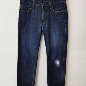 May include: Dark wash denim jeans with a classic five-pocket design. The jeans are hanging on a wooden hanger against a white background. There is a light-colored patch on the right leg.